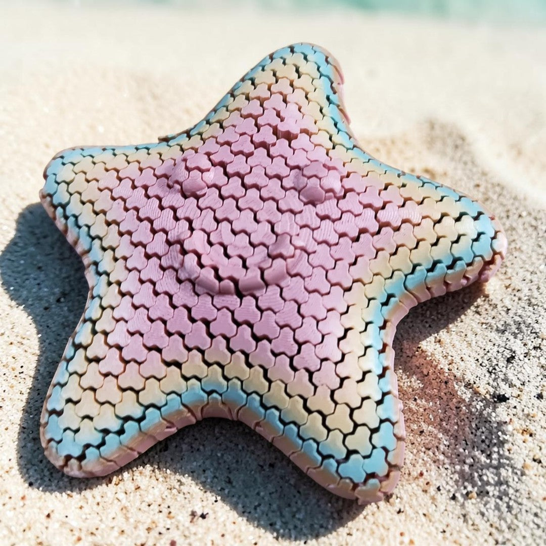 ASmaker3D articulated smiling starfish fidget toy with a pastel gradient finish resting on sandy beach sand.